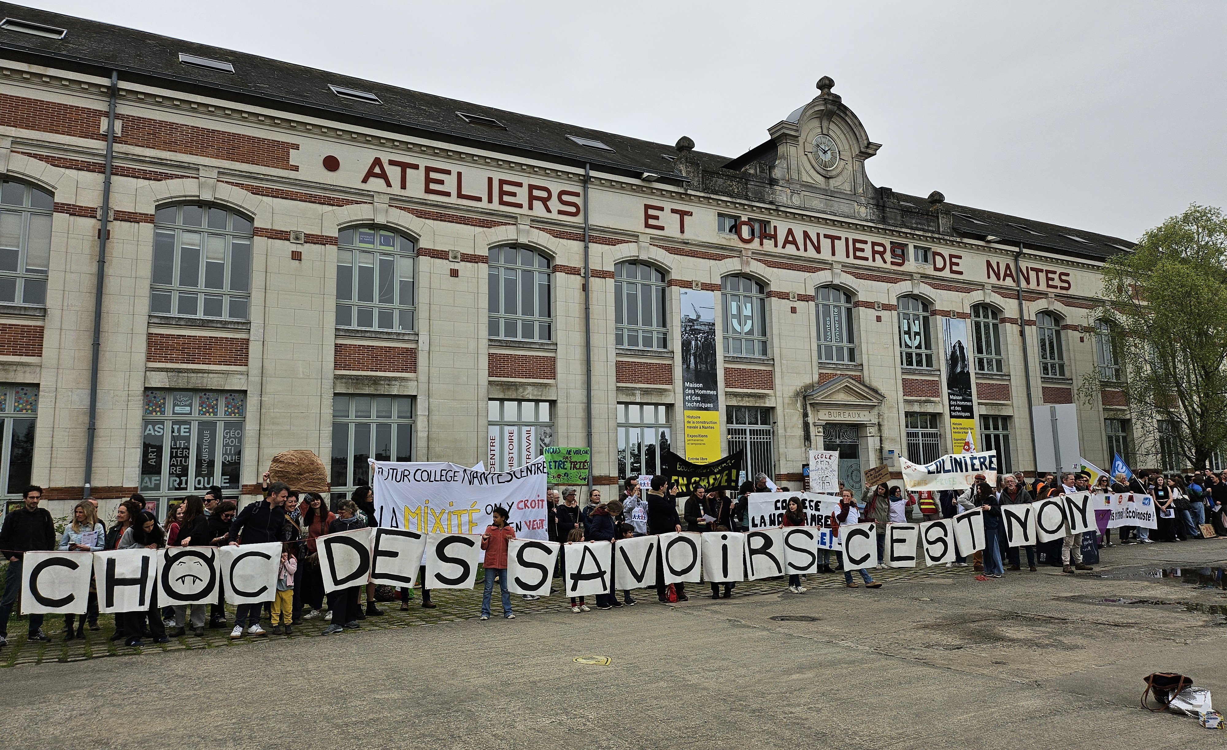 Photo d'une chaîne humaine devant le batiment Atelier et Chantiers de Nantes, les personnes portent des pancartes avec des lettres formant la phrase «Choc des savoirs c'est non» ainsi que d'autres banderoles