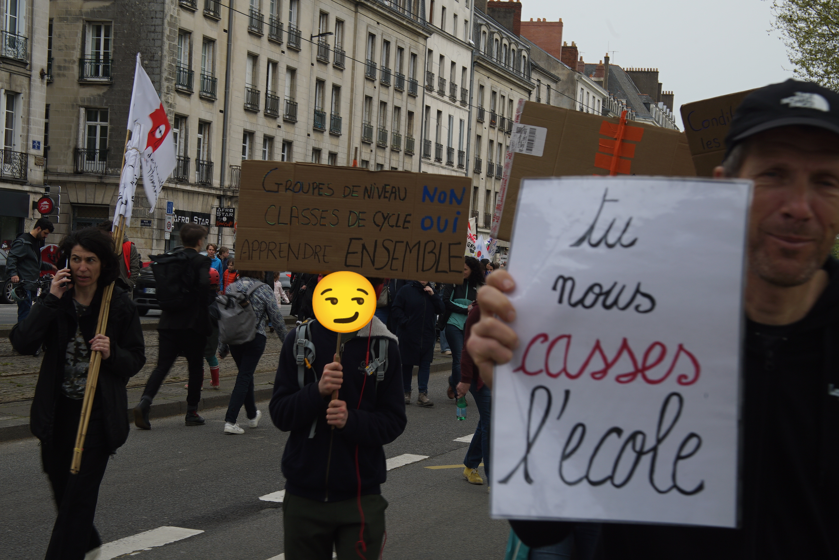 Photo de manifestants dans la rue, un adolescent au visage masqué par un smiley porte une pancarte «Groupe de niveau: Non, Classe de cycle: Oui, Apprendre ensemble. Et adulte porte une pancarte où il est indiqué «Tu nous casses l'école»